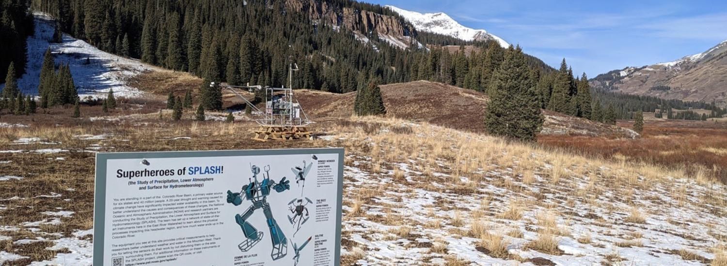 Photo of a field with snow, evergreen trees and hills. In the foreground is a large sign titled Superheroes of SPLASH and showing images of robots with labels and descriptions. In the background behind the sign is a large scientifc instrument.