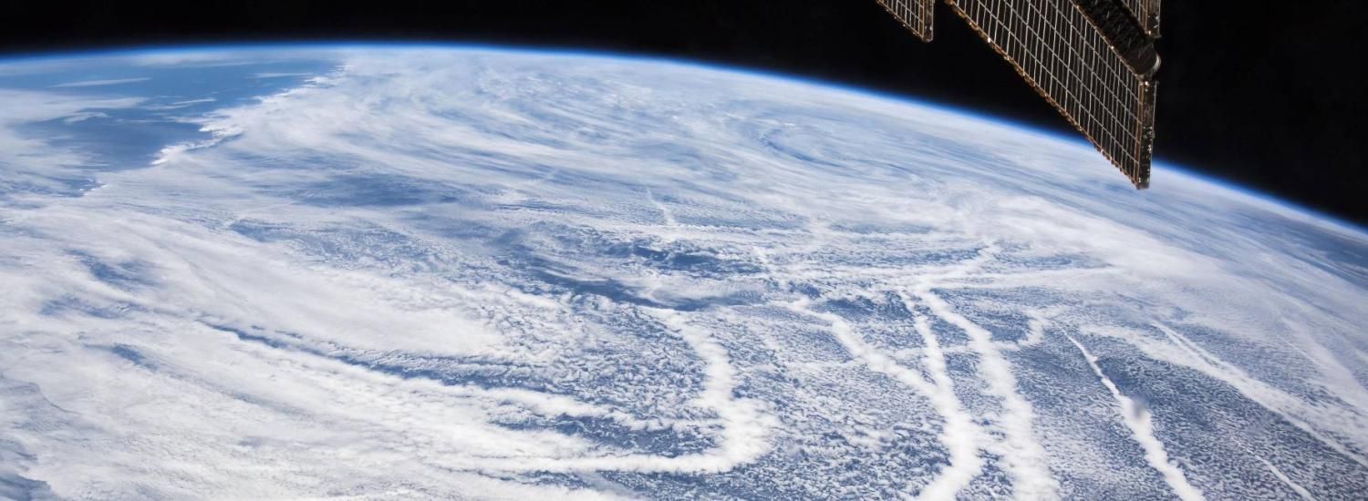 A photo of ship tracks stretching across the North Pacific Ocean near the Aleutian Islands, as seen from the International Space Station, with a solar panel of the ISS in the top right of the photograph.