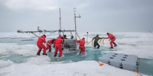 Eight scientists circled around a large instrument, moving it across melting ice, Two people are pulling the instrument and the other six are helping push the instrument. 