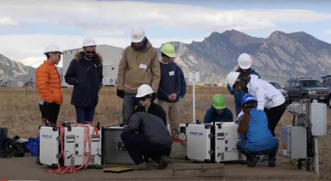 People in hard hats standing and kneeling around scientific equipment which are large white cubes with the label 'WINDCUBE'. In the background there is an open field and mountain ridge.
