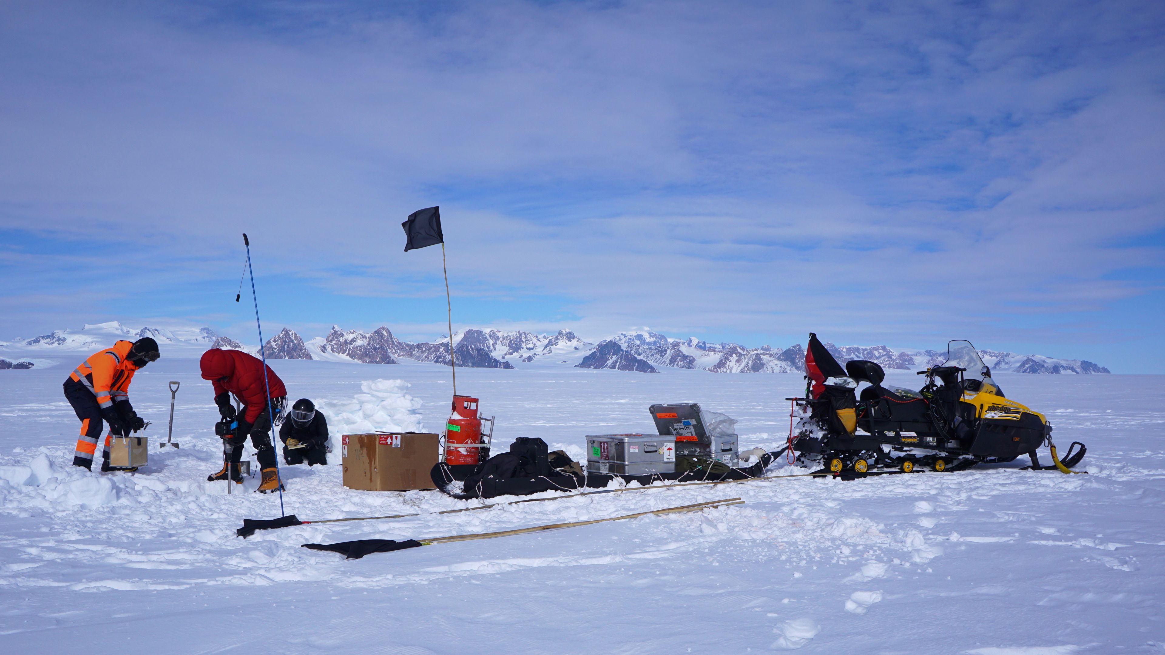 Three scientists installing instruments in the ice and snow next to their equipment and a snowmobile.