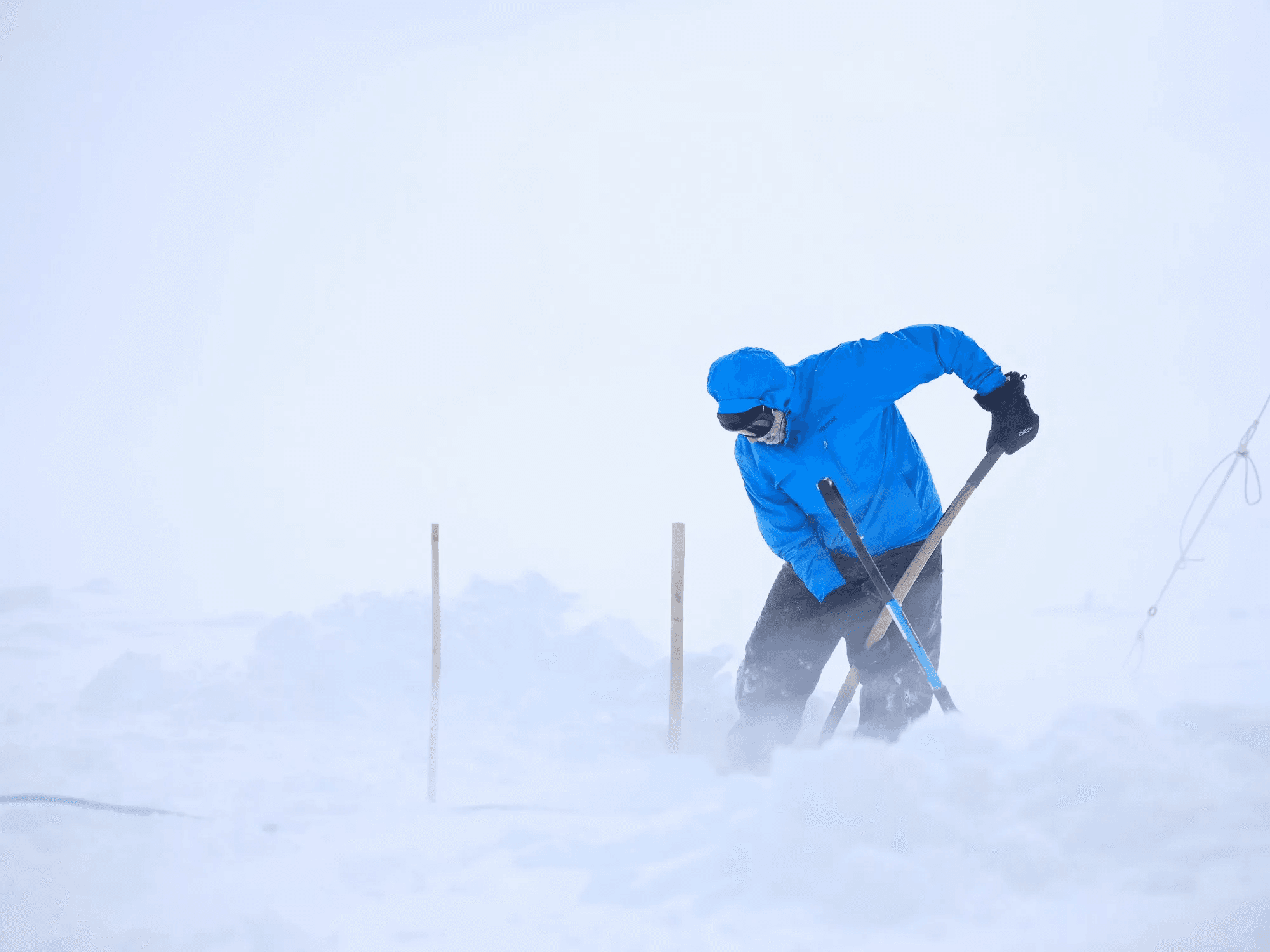 A person in cold weather gear digs a hole in blowing snow.