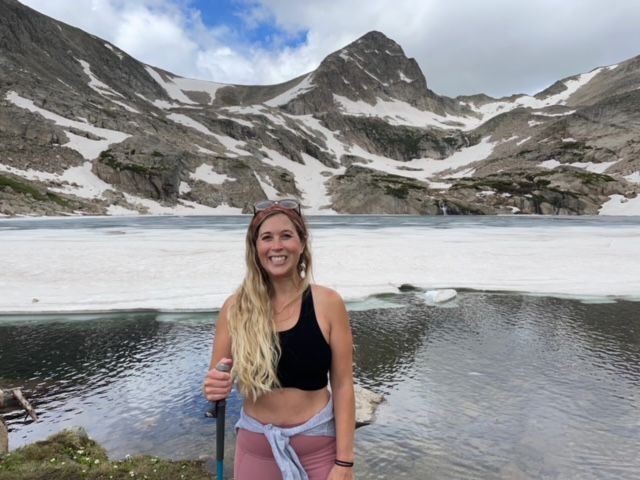 A person in hiking gear with long blonde hair poses with a hiking pole in front of a mountain lake.