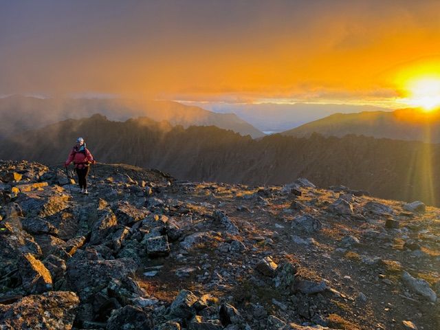 A person hikes along a rocky ridge under low clouds and a rising sun.