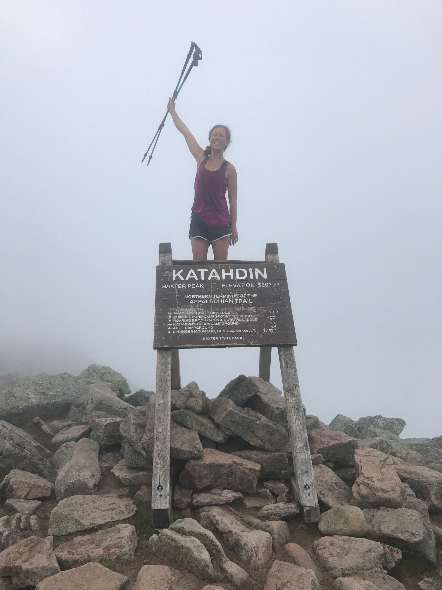 A person in a tank top and shorts stands behind a "Katahdin" sign while holding to hiking poles up in the air.