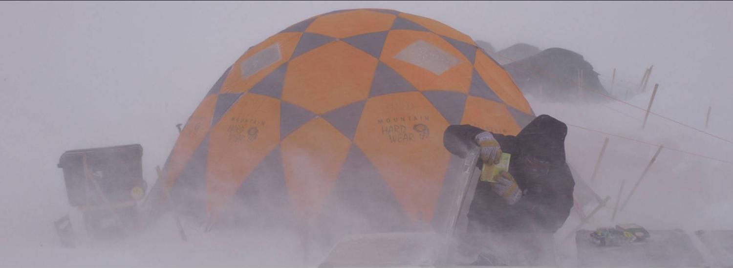 Person in jacket and globes in snow storm in front of orange tent