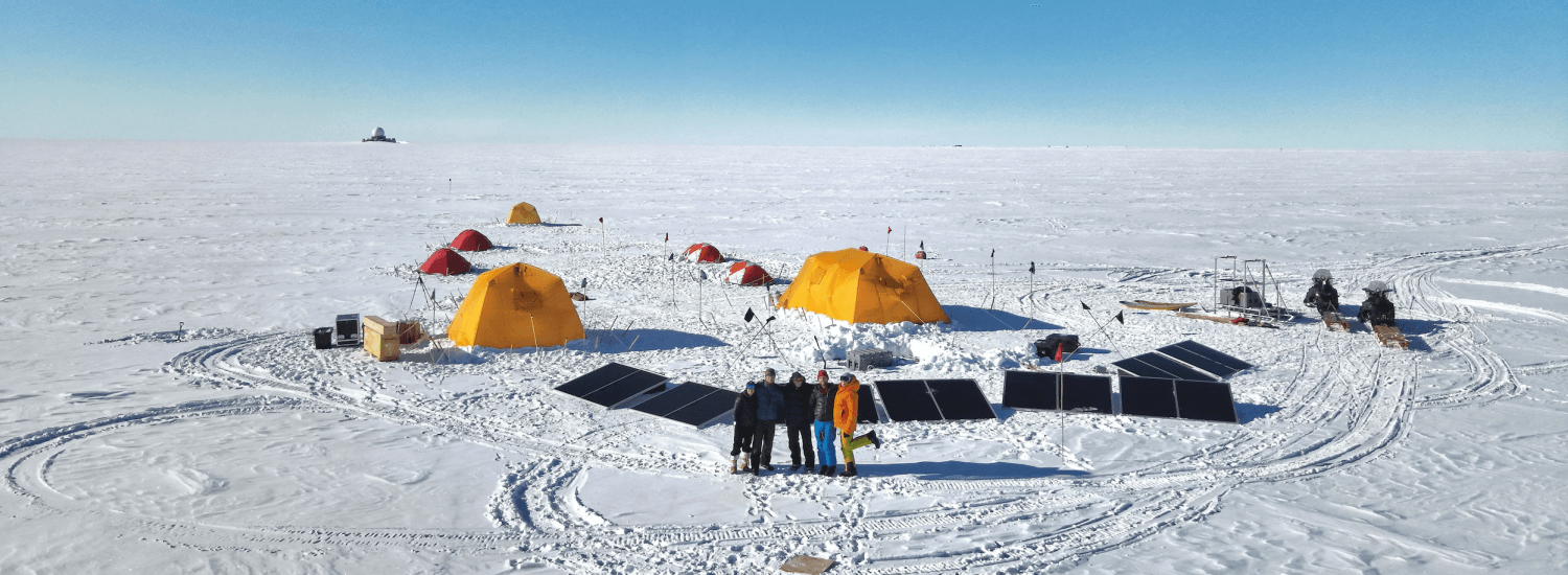Five people standing on ice and snow in front of a camp with tends, solar panels, and other equipment.