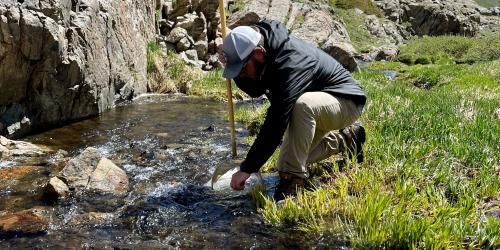 A person wearing a hat takes a water sample from a stream