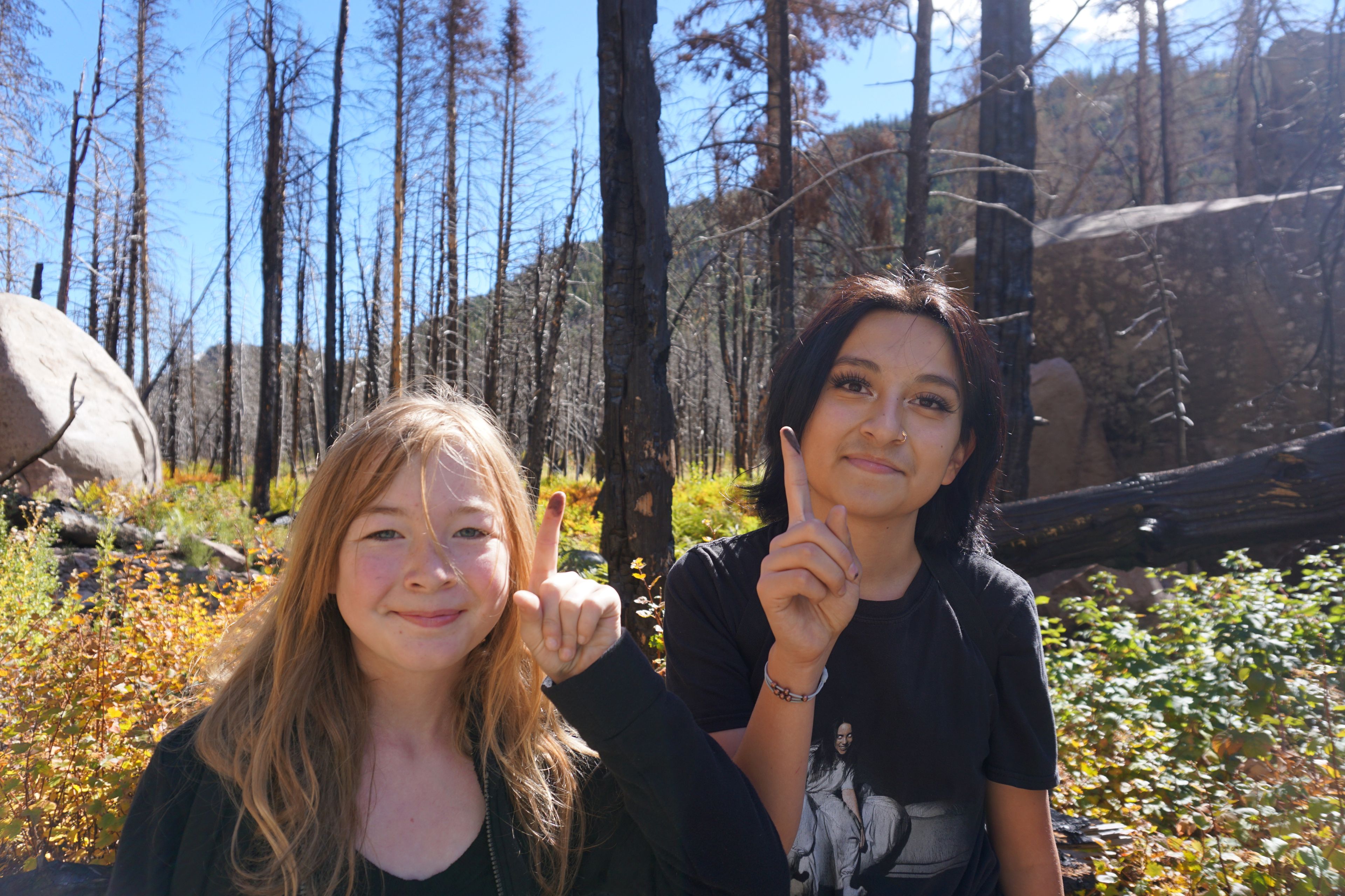 Two students stand in a stand of burned aspen trees admiring the charcoal on their fingers