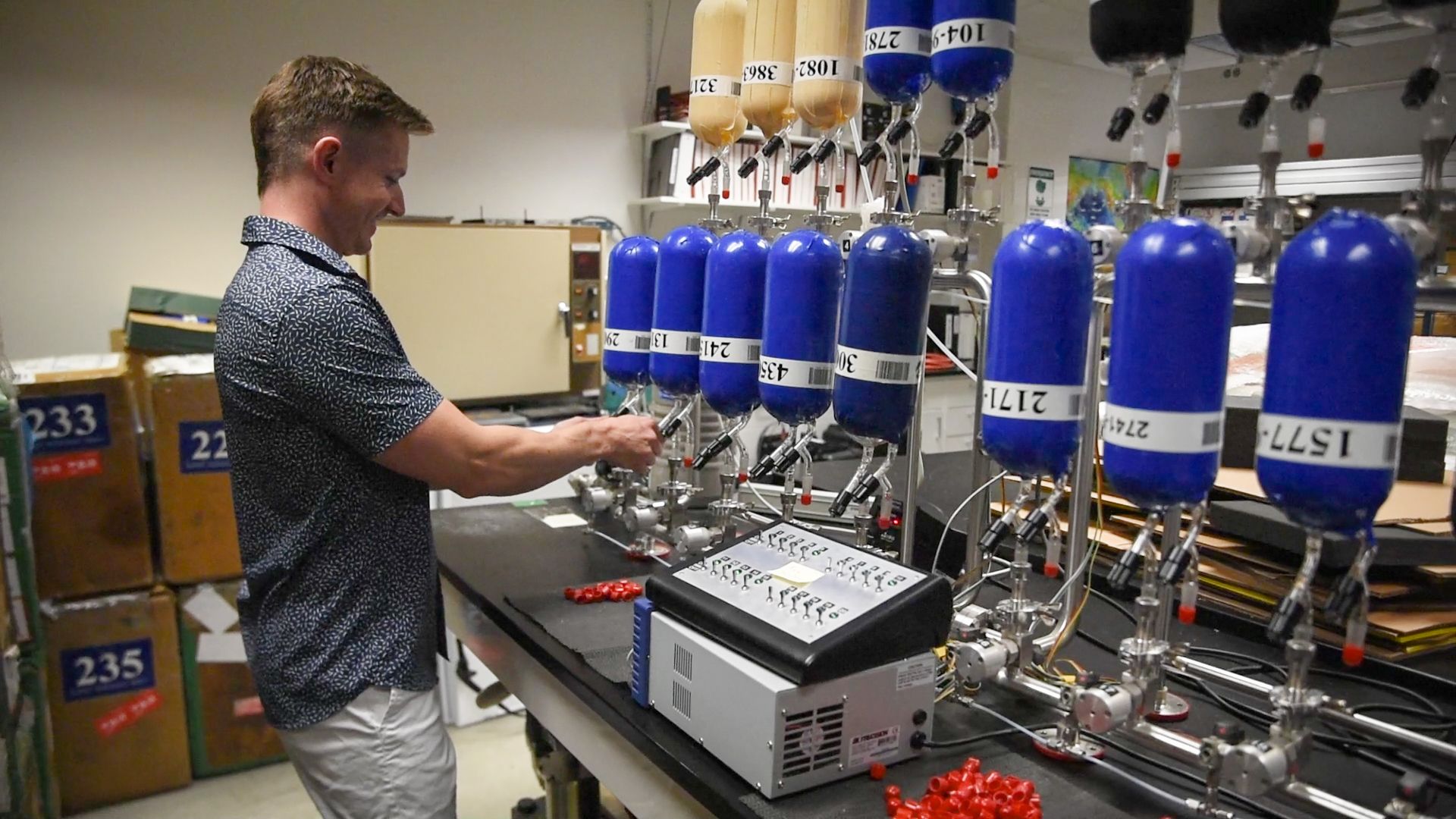 A man stands in a lab, in front of equipment with lots of blue flasks.