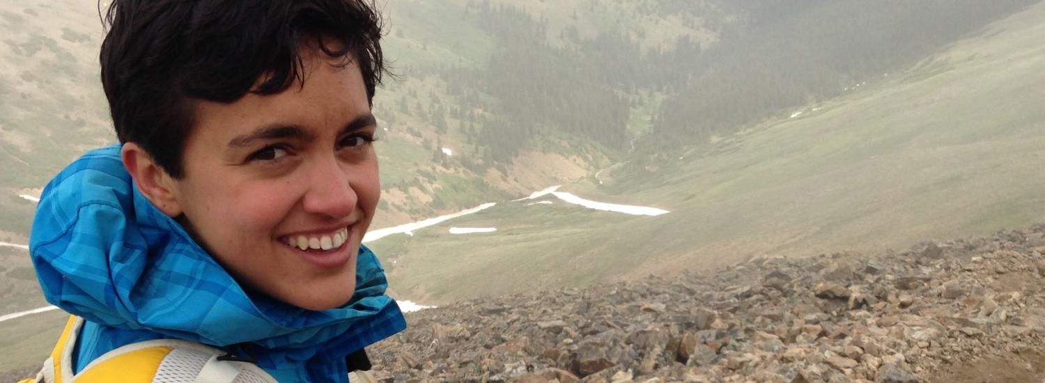 Young person smiles at the camera above a scree field into a mountain valley