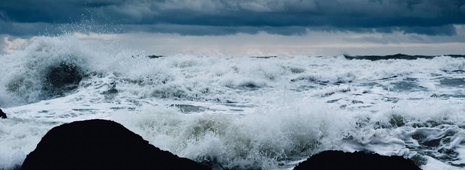 White-capped waves crashing along a dark, rocky shore with dark storm clouds in the horizon.