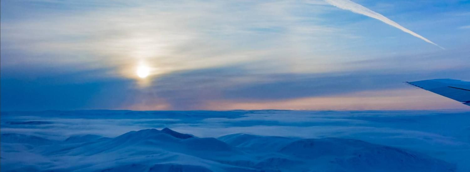 Photo looking out of a NASA DC-8 flying over the Canadian Arctic, with a view of the sun, clouds, and icy mountains. 