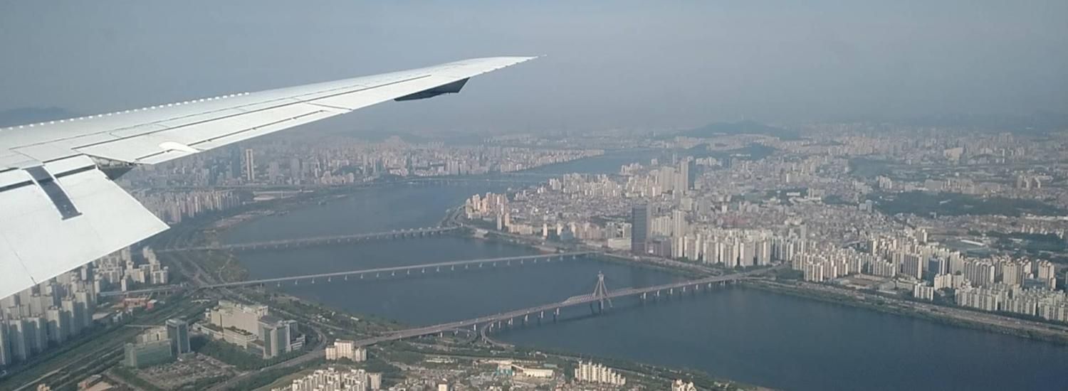 A hazy sky, city, and river, as seen by air with an airplane wing in frame