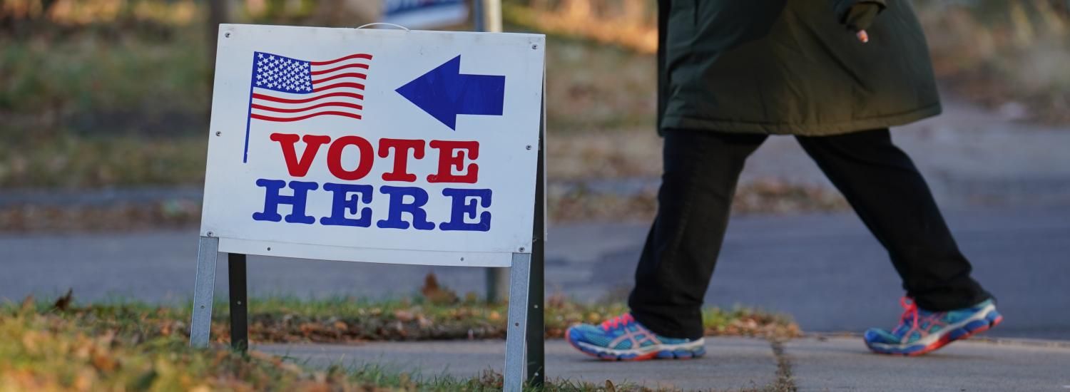 A voter in sneakers walks by a sidewalk sign saying vote here in the fall
