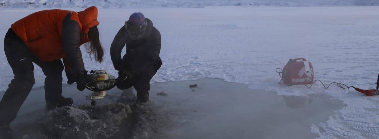 Two researchers use an augur to drill into ice atop a frozen river