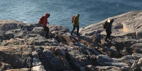Three hikers with backpacks walk on a rocky shore along water's edge.