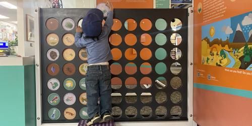A young child stands on a step stool and reaches above the Connect 4 board to insert a game token.