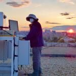 Researcher adjusts equipment on an urban rooftop as the sun sets in the background