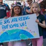 Two girls standing side by side holding a sign that reads "The seas are rising but so are we!"