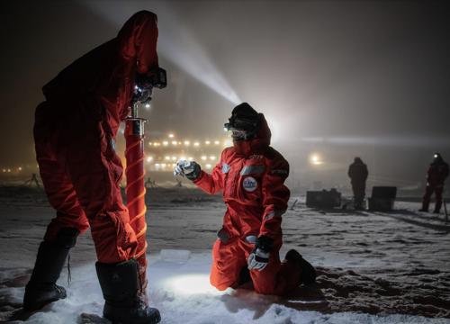 MOSAiC scientists drill through the Arctic ice with the research vessel Polarstern illuminated in the background.
