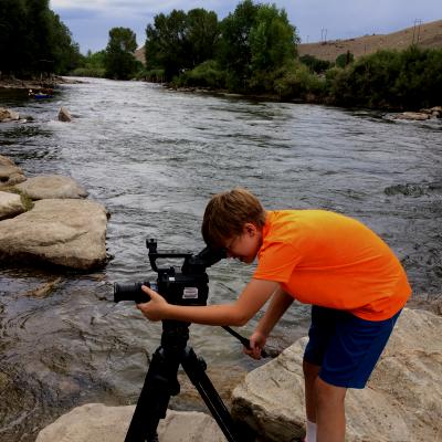 A boy uses a video camera in front of a river.