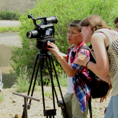 A mentor helps a child line up a shot on a video camera.