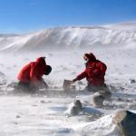Two scientists in large red parkas kneeling on the ground working on scientific equipment in a snowy landscape while a cold wind whips snow up around them.
