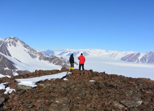 Two scientists stand on a rocky outcrop looking over a field of snow ringed with mountains.