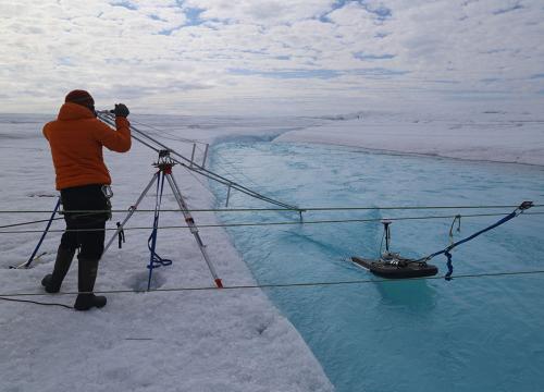 A scientist is an orange coat uses a scientific instrument to measure rates of a glacial floe on a stark white glacier that disappears into white clouds.