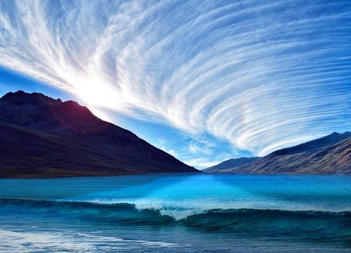Whispy swirling clouds over a body of water ringed with mountains.