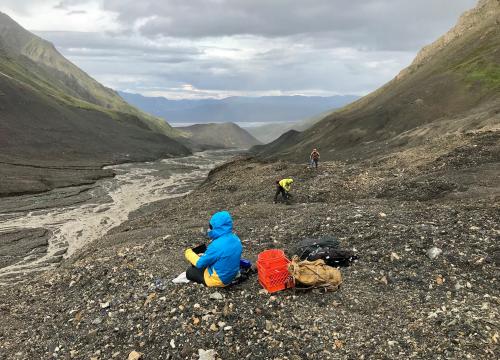 A scientist sits in a field of rubble where a glacier used to be while two additional scientists collect samples in the background.