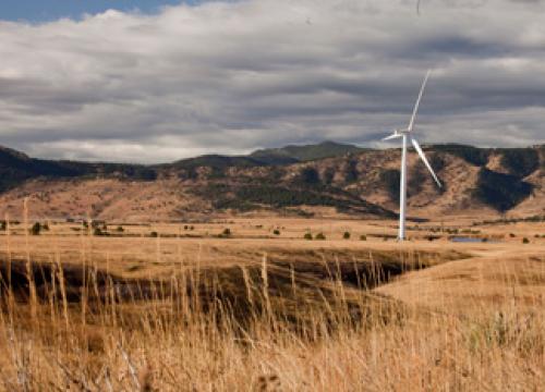 A white wind turbine turns in front of green and tan mountains. There are dried tall grasses in the foreground.
