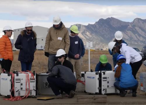 People in hard hats standing and kneeling around scientific equipment which are large white cubes with the label 'WINDCUBE'. In the background there is an open field and mountain ridge.