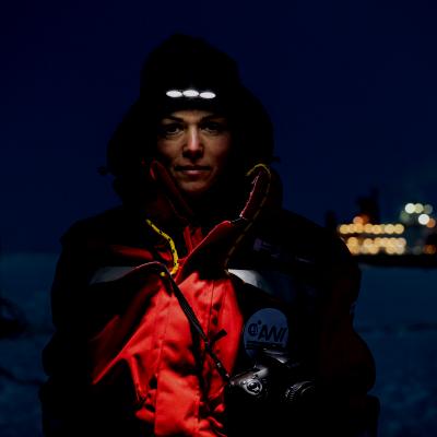A scientist illuminated by their own headlamp in darkness with just the lights of a distant ship behind her.