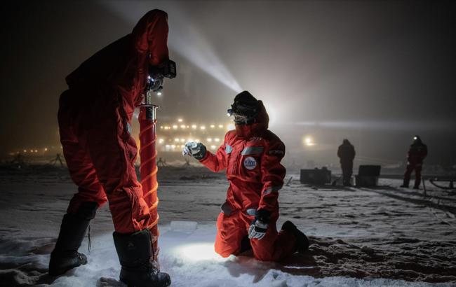 MOSAiC scientists drill through the Arctic ice with the research vessel Polarstern illuminated in the background.